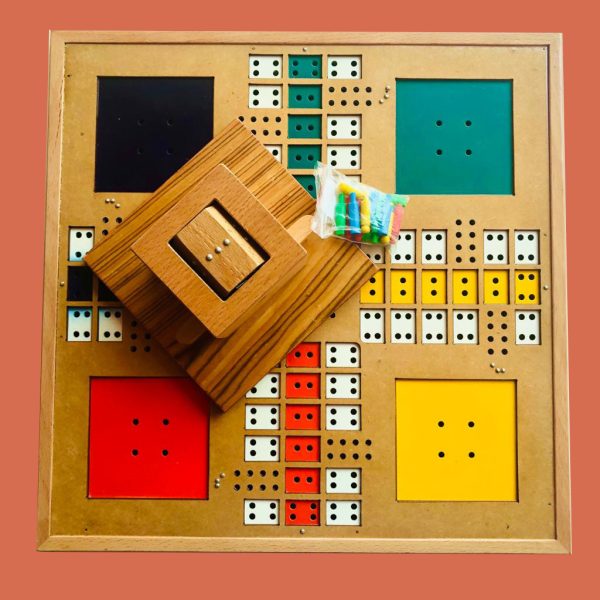 La imagen muestra un tablero de juego de ludo. El tablero es cuadrado y está dividido en cuatro secciones de colores: negro, verde, rojo y amarillo. Cada sección tiene un área cuadrada grande y varias casillas pequeñas con puntos, que representan las casillas del juego. En el centro del tablero hay una estructura de madera que parece ser una caja o soporte, y sobre ella hay una pequeña bolsa de plástico que contiene fichas de colores
